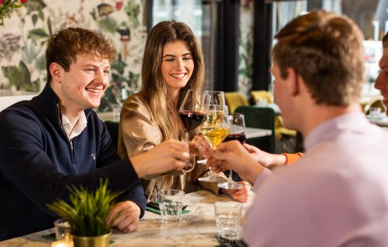 Een vrouw en twee mannen van in de twintig zitten aan tafel bij Loev Hengelo en proosten met hun drankjes.